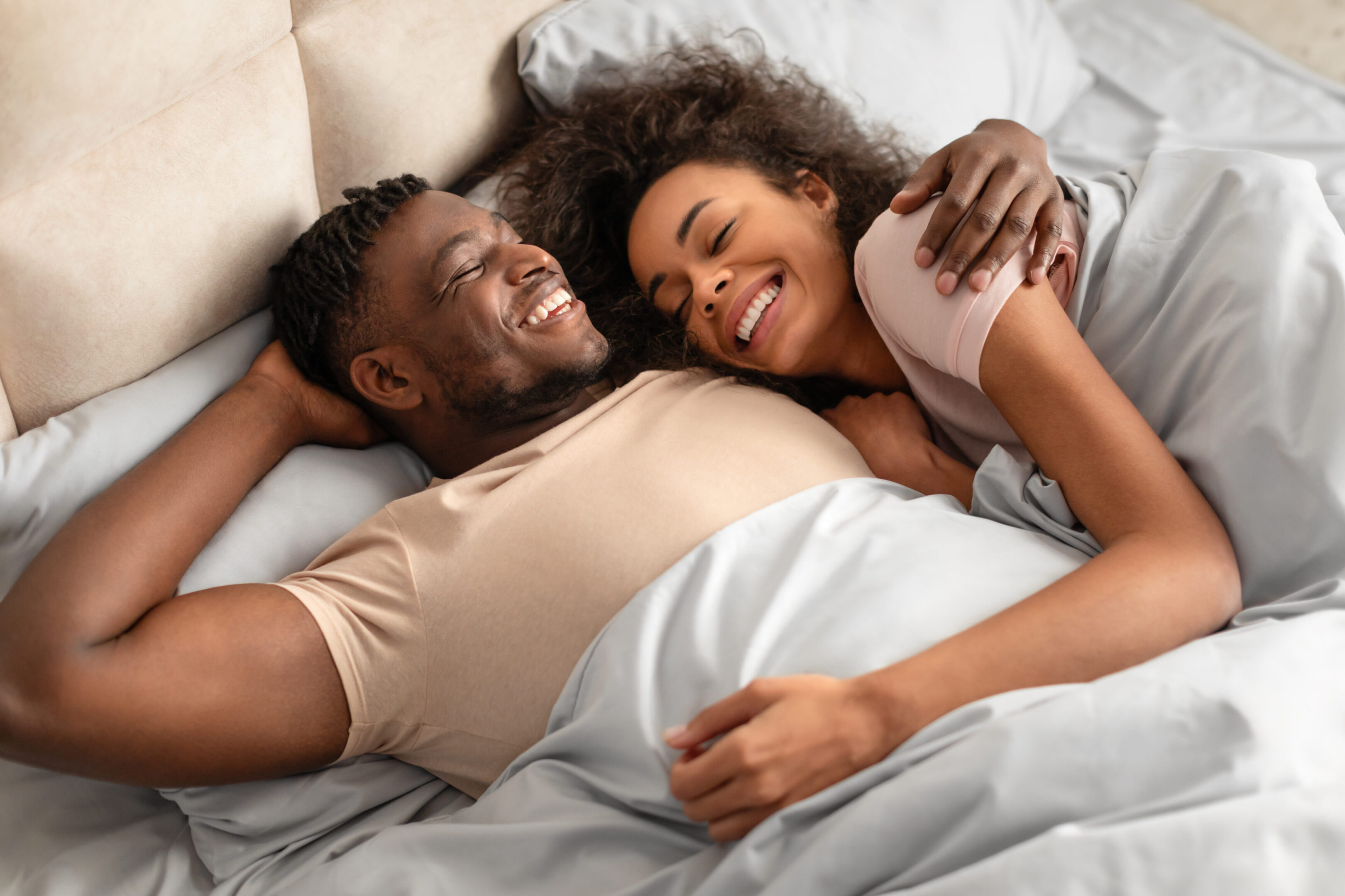 black man and woman lying in bed hugging radiating happiness A man and woman laying in bed smiling.
